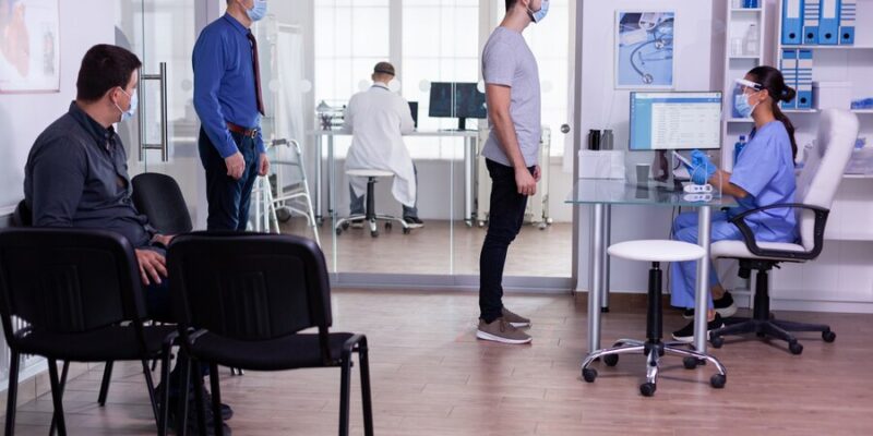 young-man-checking-appointment-respecting-social-distancing-hospital-waiting-room-nurse-looking-computer-wearing-face-mask