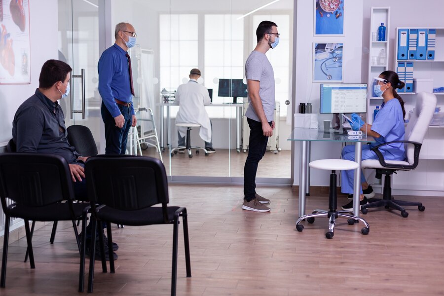 young-man-checking-appointment-respecting-social-distancing-hospital-waiting-room-nurse-looking-computer-wearing-face-mask